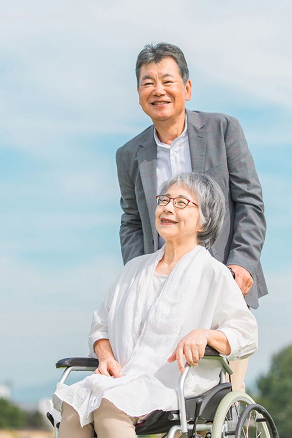 車椅子の高齢女性と寄り添う男性が青空の下で笑顔で過ごしている様子
